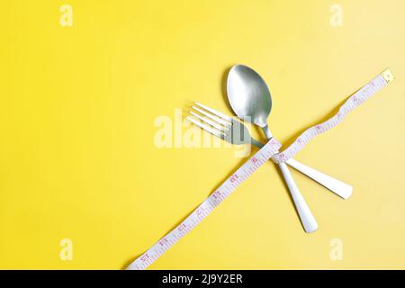 Gabel und Löffel mit Messband umwickelt flach in gelbem Hintergrund liegen. Anorexie, Ernährung und Ernährung, Gewicht zu verlieren Konzept. Stockfoto