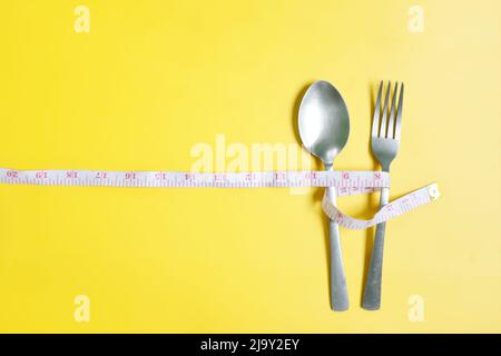 Gabel und Löffel mit Messband umwickelt flach in gelbem Hintergrund liegen. Anorexie, Ernährung und Ernährung, Gewicht zu verlieren Konzept. Stockfoto