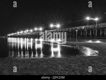 Die Lichter des Pier in der Nacht reflektierten sich im nassen Sand am Strand. Hölzerner Pier am White Rock, BC, erstreckt sich diagonal in das Bild. Schwarzweiß-Foto Stockfoto