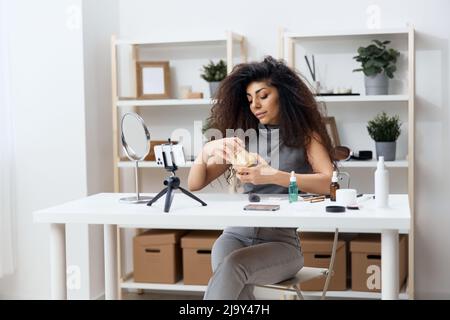 Fröhlich gebräunt schöner lockiger lateinischer Beauty-Blogger in Casual Top erzählt von Körpercreme im Heim-Interieur. Space Mockup Banner kopieren. Influencer-Rekord Stockfoto