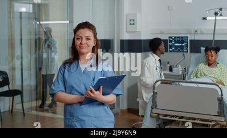 Porträt einer Frau, die als Krankenschwester in der Hand von Checkup-Papieren arbeitet, während sie auf der Krankenstation steht. Medizinische Assistentin mit Dokumenten, die auf die Kamera blicken und lächeln, und hilft dem Spezialisten. Stockfoto
