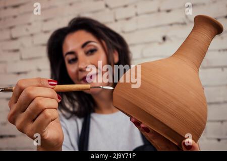 Junge und attraktive Töpferin, die in einem Atelier mit einem braunen Tonvasatopf in der Handwerkswerkstatt arbeitet und Werkzeug und Becher in der Hand hält Stockfoto