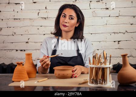 Junge und attraktive Töpferin, die in einem Atelier mit einem braunen Tonvasatopf in der Handwerkswerkstatt arbeitet und Werkzeug und Becher in der Hand hält Stockfoto