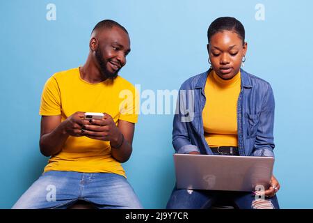 Millennials koppeln sich im Studio mit Smartphone und Laptop und haben Spaß mit Technologie auf blauem Hintergrund. Mann und Frau in Beziehung mit Blick auf Mobiltelefon und modernen Computer. Stockfoto