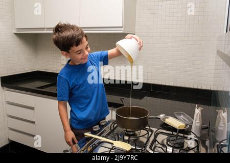 8-jähriges Kind in der Küche lächelt und gießt Kondensmilch in die Pfanne, um brigadeiro zu machen. Stockfoto