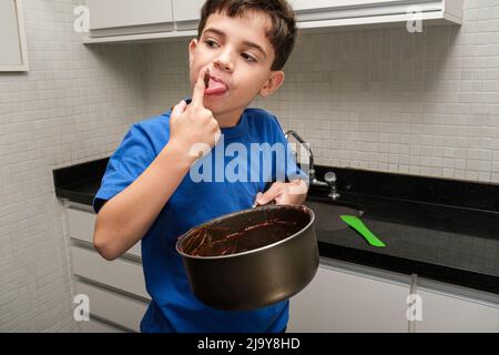 8-jähriges Kind in der Küche, das eine Pfanne in der Hand hält und mit brigadeiro (Brigadier) seinen Finger leckt. Stockfoto