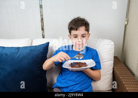 8 Jahre altes Kind sitzt auf dem Sofa und schaut mit dem süßen brigadeiro auf den Löffel. Stockfoto