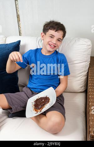 Lächelndes 8-jähriges Kind, das auf dem Sofa sitzt, einen Löffel mit dem süßen brigadeiro hält und auf die Kamera schaut. Stockfoto