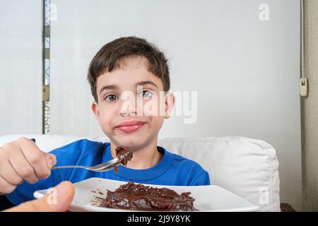 8-jähriges Kind hält einen Löffel mit süßem brigadeiro und schaut mit einem lustigen Gesicht auf die Kamera. Stockfoto