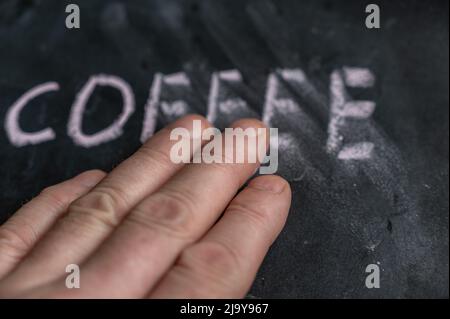 Das halbgelöschte Wort KAFFEE auf der schwarzen Tafel. Die linke Hand eines erwachsenen Mannes entfernt das handgeschriebene Wort mit seinen Fingern. Unscharfe Buchstaben auf schwarzem Su Stockfoto