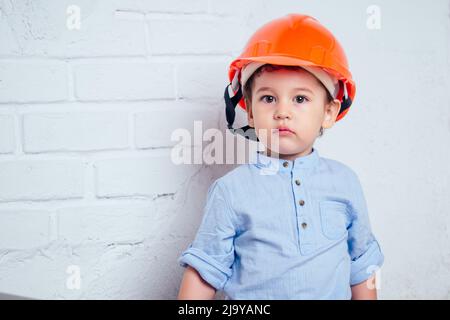 Junge Baumeister halten Bürstenrolle charmant niedlichen kleinen Jungen in Helm Hardhat bemalte Wand mit Farbe weiß Farbe. Konzept der Reparaturen Hauswohnung Stockfoto