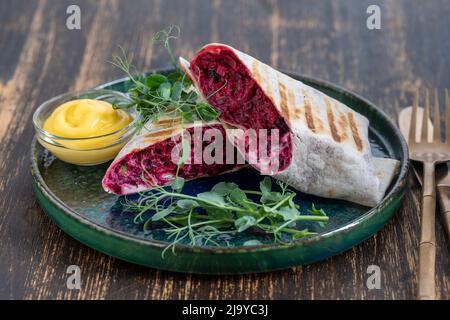 Veganer Burrito. Aufgeschnittene rohe Lebensmittelverpackung mit roter Rote Beete, Feta-Käse, Pflaumen, Dill und Rahmsoße mit Erbsen-Mikrogemüse auf einem Teller. Nahaufnahme Stockfoto