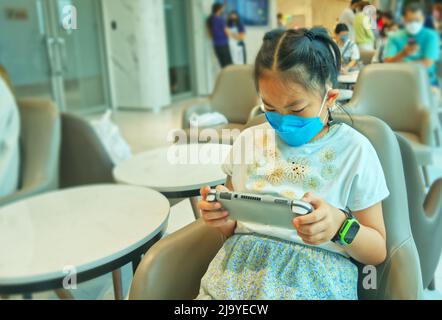 Portrait Asian Kind Mädchen spielt ein tragbares Spiel Handheld-Konsole, während auf einen Arzt in einem Krankenhaus, trägt medizinische Gesichtsmaske, verschwommen Bac Stockfoto