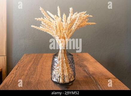 Einfache Komposition mit Glasvase und trockenem Bouquet mit Ähren aus Weizen auf Holztisch, Stillleben mit dem sonnigen Licht Stockfoto