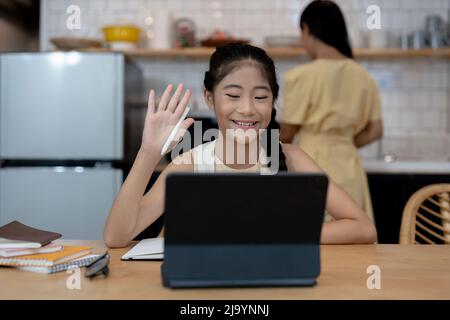 Lächelndes kleines asiatisches Mädchen in Kopfhörern haben Videoanruf entfernte Klasse mit Lehrer mit Laptop, glückliche kleine Kind Welle Gruß mit Tutor, Studie Stockfoto