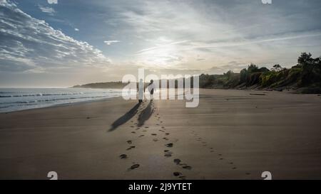 Pärchen, die am Strand spazieren gehen. Die Sonne scheint durch die Wolken, New Plymouth. Stockfoto