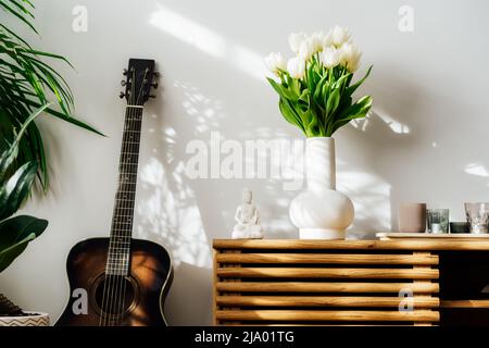 Skandinavische minimalistische Einrichtungsdetails - Bouquet von weißen Tulpenblumen in Vase, Kerzen, Buddha-Statuette auf Holzschrank. Grünhausanlage Stockfoto