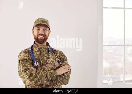 Glücklicher Armeearzt in Tarnuniform, der mit gekreuzten Armen und lächelnd stand Stockfoto