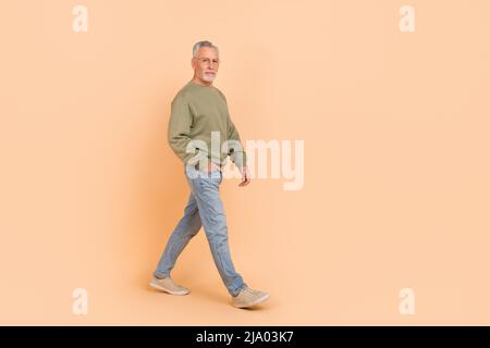 In voller Länge Foto von funky alte graue Frisur Mann gehen tragen Brillen Pullover Jeans Sneakers isoliert auf beigem Hintergrund Stockfoto