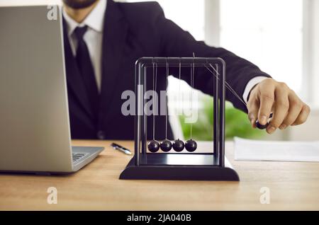 Geschäftsmann, der mit einem Laptop am Schreibtisch sitzt und mit Newtons Pendel spielt Stockfoto