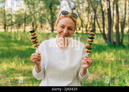 Reife Frau 50 Jahre alt Essen gegrilltes grillgemüse von Holzspießen im Freien im Sommerwald Stockfoto