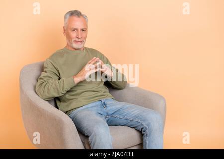 Foto von strengen Holder grau Frisur Mann sitzen tragen Pullover Jeans isoliert auf beigem Hintergrund Stockfoto