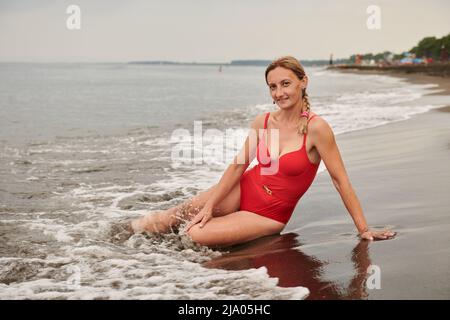 Brunette in einem roten Badeanzug liegt am Strand am Meer Stockfoto