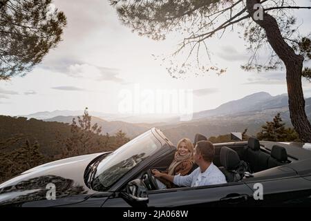 Junges, fröhliches Familienpaar auf einer Fahrt mit einem Cabriolet zwischen den Bergen Stockfoto