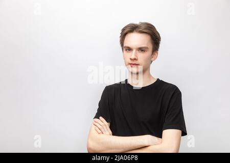 Porträt eines verdächtigen jungen Mannes mit schwarzem T-Shirt, der seine Hände gekreuzt hielt und auf die Kamera schaute. Studio auf grauem Hintergrund aufgenommen Stockfoto