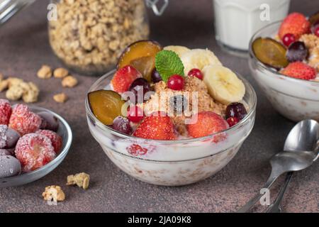 Müsli knuspriges Müsli mit Naturjoghurt, gefrorenen Beeren und Früchten und Nüssen in zwei Glasschüsseln auf braunem Hintergrund, gesundes Frühstück Stockfoto