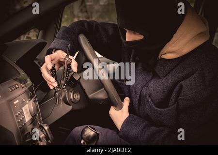 Ein Autodieb versucht, das Zündschloss mit einem Werkzeug zu öffnen. Der Autodieb versteckte sein Gesicht in einer Sturmhaube Stockfoto