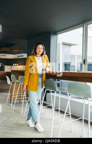 Fröhliche Geschäftsfrau, die mit einem Smartphone in der Hand auf die Kamera schaut. Ethnische Geschäftsfrau, die aus der Ferne in einem Co-Working-Bereich arbeitet Stockfoto
