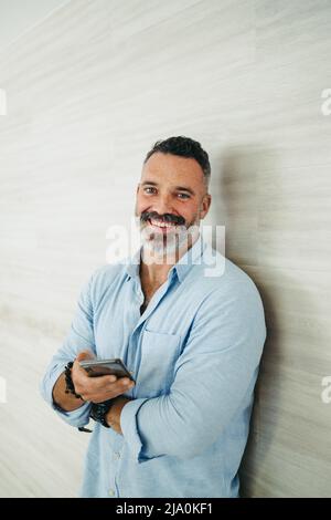 Fröhlicher Unternehmer, der die Kamera anlächelt, während er ein Smartphone hält und gegen eine Wand steht. Glücklicher, reifer Geschäftsmann, der eine Textnachricht einsendet Stockfoto