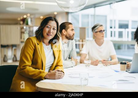 Fröhliche Geschäftsfrau, die bei einem Treffen mit ihren Kollegen auf die Kamera schaut. Gruppe von innovativen Design-Profis Austausch kreativ Stockfoto