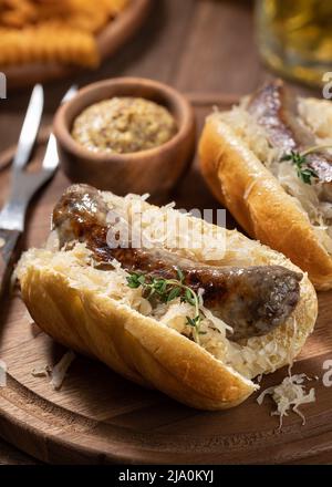 Bratwurst mit Sauerkraut auf einem Brötchen und Senf in einer Schüssel auf einem hölzernen Schneidebrett Stockfoto