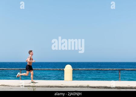 Ein Jogger oder Läufer, der hart an einer Straße am Meer trainiert und bei heißem Sonnenschein bis zur Taille entkleidet ist. Im Hintergrund ist ein türkisfarbenes Meer zu sehen Stockfoto