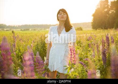 Schöne Frau in weißem Sonnenkleid, die die Sommerlandschaft genießt. Pflücken Sie farbenfrohe Blumen, atmen Sie frische Luft und blumigen Duft ein, das sonnige Feld Stockfoto