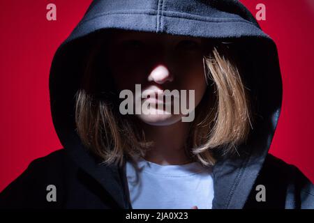 Ein Mädchen im Teenageralter in einem schwarzen Hoodie mit einer Kapuze auf rotem Hintergrund. Das Gesicht ist geschlossen. Das Konzept der Probleme bei Jugendlichen und die Schwierigkeiten der t Stockfoto
