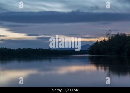 Landschaft aus Fluss und Berg Silhouette in der Dämmerung, Sava Fluss mit bewaldeten Ufer und Motajica Bergszene mit Wolken am Himmel während der blauen Stunde Stockfoto