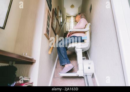 Ältere Frau, die einen automatischen Treppenlift auf einer Treppe in ihrem Haus verwendet. Medizinischer Treppenlift für behinderte und ältere Menschen im Haus. Selektive Stockfoto