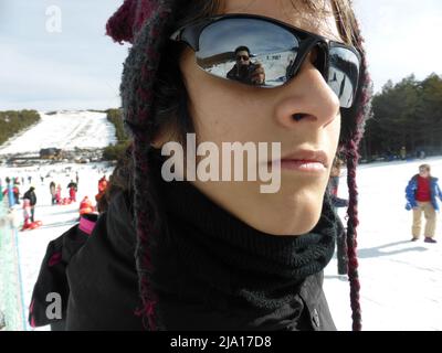 Nahaufnahme eines Jungen im Schnee, der einen Wollhut und eine Sonnenbrille mit verspiegelten Gläsern trägt. Der Fotograf spiegelt sich im Glas des Glases wider Stockfoto