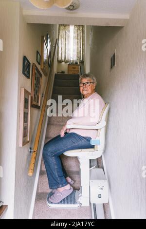Ältere Frau, die einen automatischen Treppenlift auf einer Treppe in ihrem Haus verwendet. Medizinischer Treppenlift für behinderte und ältere Menschen im Haus. Vertikales Fahrzeug Stockfoto