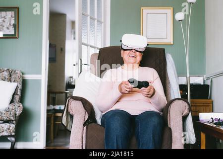 Emotionale Senior-Frau trägt Virtual-Reality-Brille und mit Wireless-Controller, Joystick Gamepad spielen Videospiele zu Hause. Spaß im Ruhestand. Stockfoto