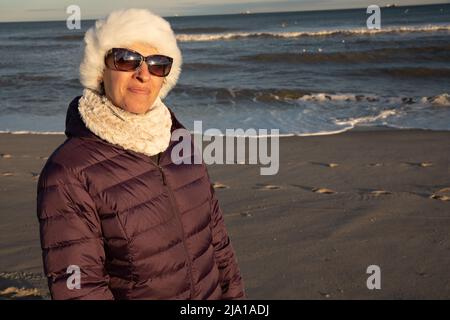 Frau Ende 40s am Strand in einem weißen Hut und einer lila Jacke im Winter Stockfoto