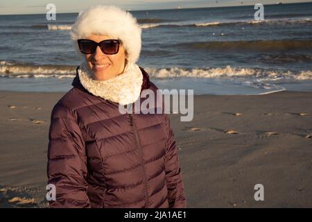 Frau Ende 40s am Strand in einem weißen Hut und einer lila Jacke im Winter Stockfoto