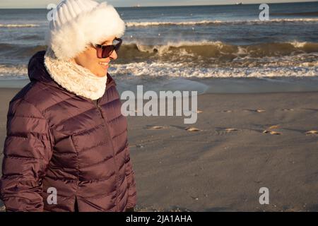 Frau Ende 40s am Strand in einem weißen Hut und einer lila Jacke im Winter Stockfoto