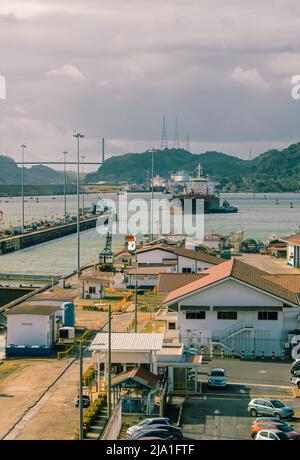 Ein Frachtschiff am Panamakanal, das durch die Schleusen des Kanals fährt, mit Hafeninfrastruktur, Instandhaltungsgebäuden und Kontrolleinrichtungen im Blick. Stockfoto