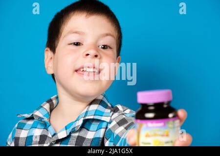 Ein 4-jähriger Junge hält ein Glas Vitamin D oder andere Nahrungsergänzungsmittel auf blauem Hintergrund in der Hand. 22. Mai 2022, Zaporozhye, Ukraine. Stockfoto