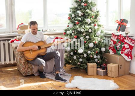 Glücklicher junger Mann spielt Gitarre. Guy sieht glücklich und sorglos aus. Mann in festlichem Hut allein feiern Weihnachten oder Neujahr. Weihnachtsbaum Stockfoto