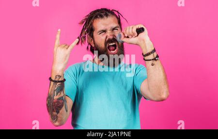 Bärtiger Mann, der im Mikrofon mit Hörnern singt, ist eine Geste eines Heavy Metal Rock. Hipster genießen Karaoke. Stockfoto